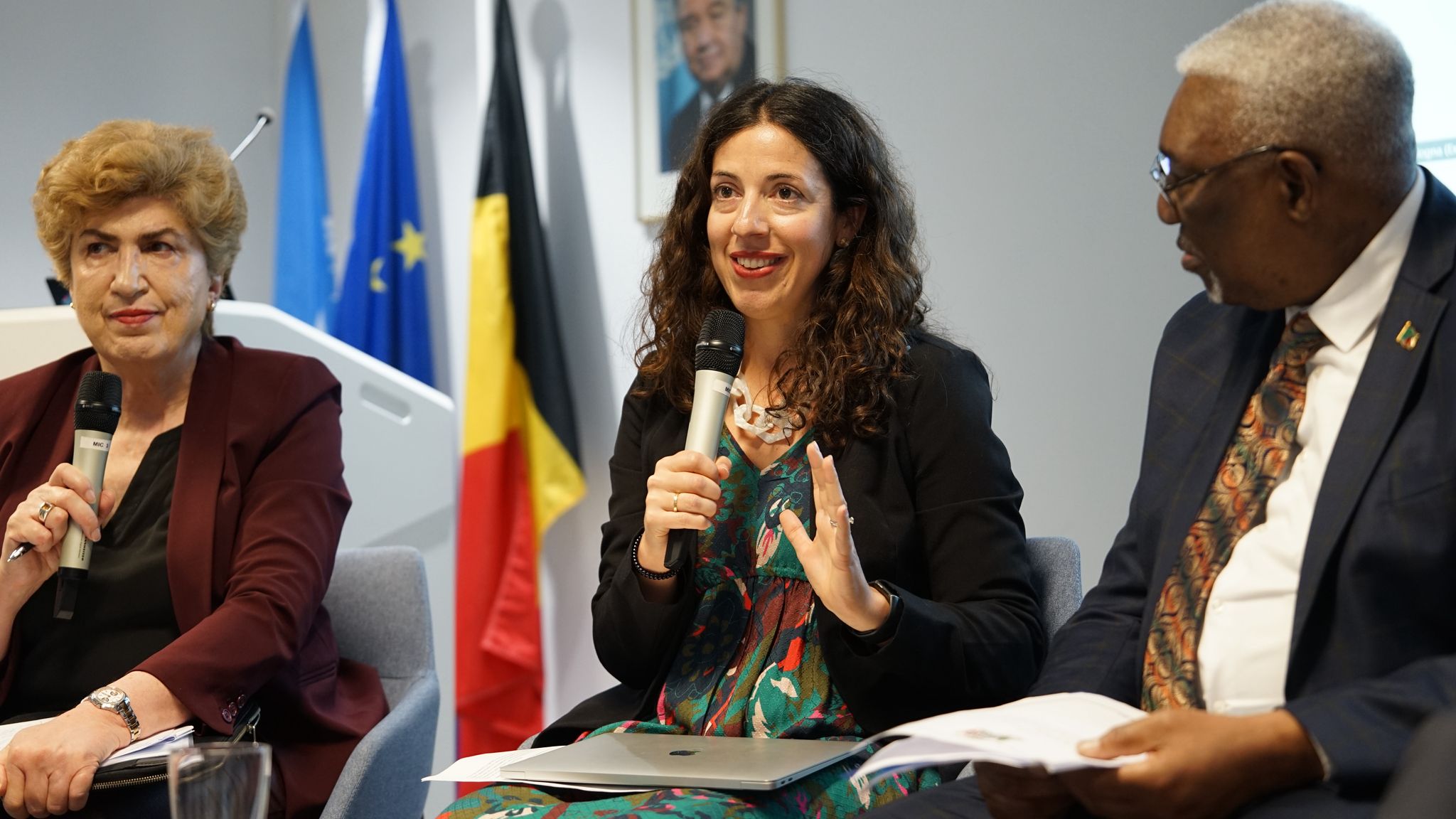 Dr. Maria Ron Balsera, CESR’s Executive Director, speaks during a panel discussion, holding a microphone and gesturing with her hand. She is seated between two other panelists, with international flags visible in the background.